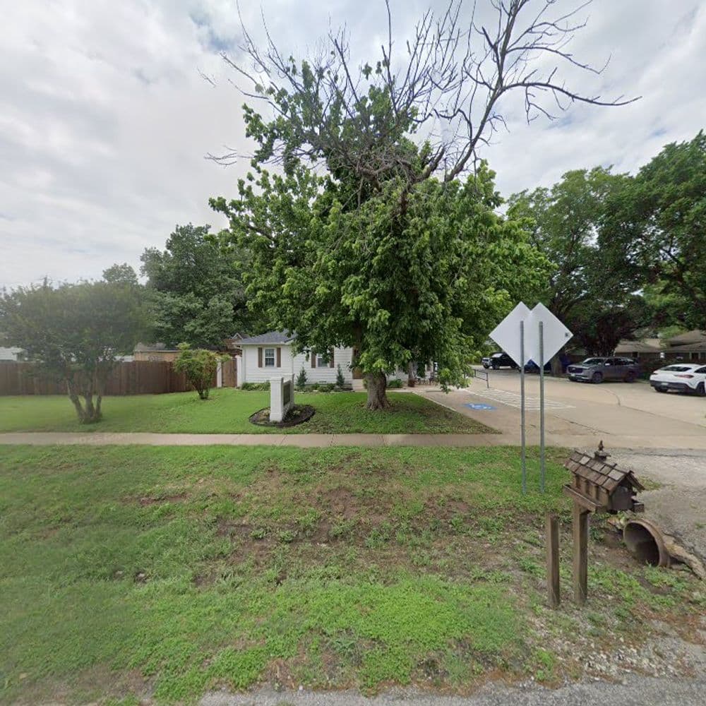 Home exterior with large tree, green grass, and parked cars nearby on a residential street.