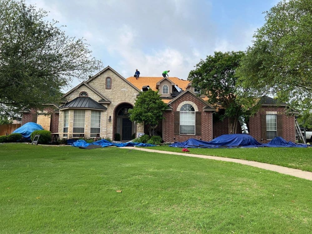 Home undergoing roof repairs with workers and blue tarps on a sunny day.