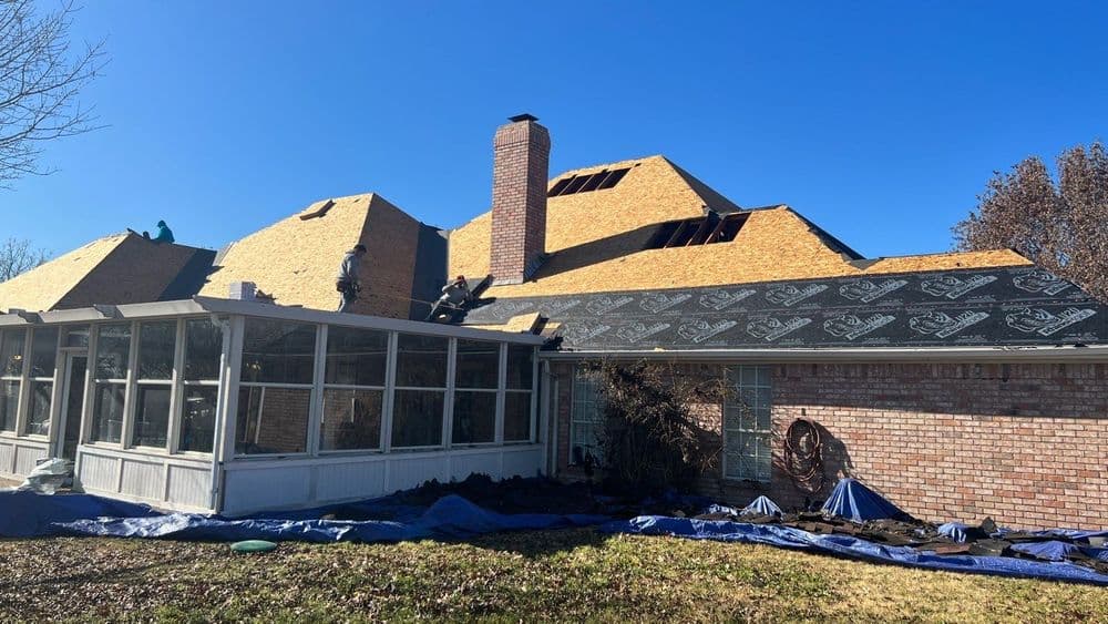 Roofing contractors installing shingles on a house with a sunroom and clear blue sky.