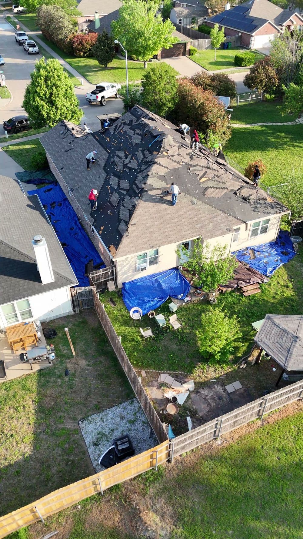 Workers replacing a roof, covering with black shingles, surrounded by residential homes and greenery.
