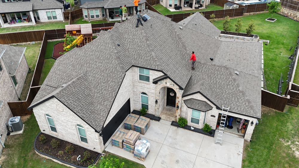 Roofing workers installing shingles on a residential home with patio and landscaping visible.