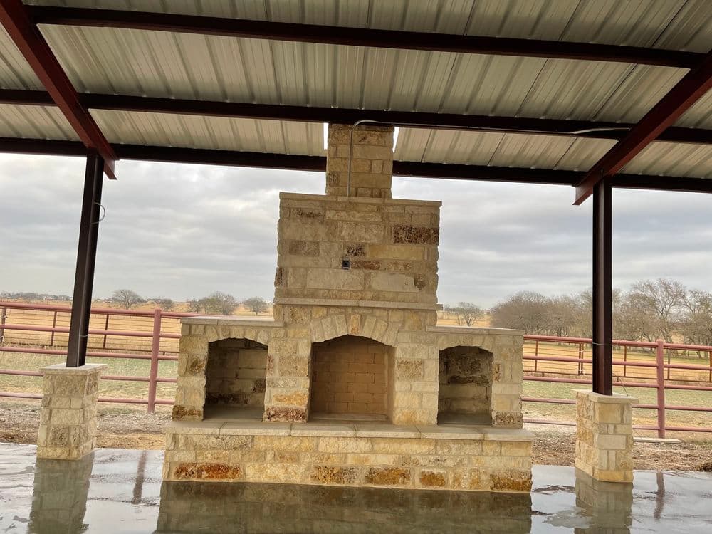 Stone outdoor fireplace under metal roof with open storage areas, surrounded by scenic landscape.