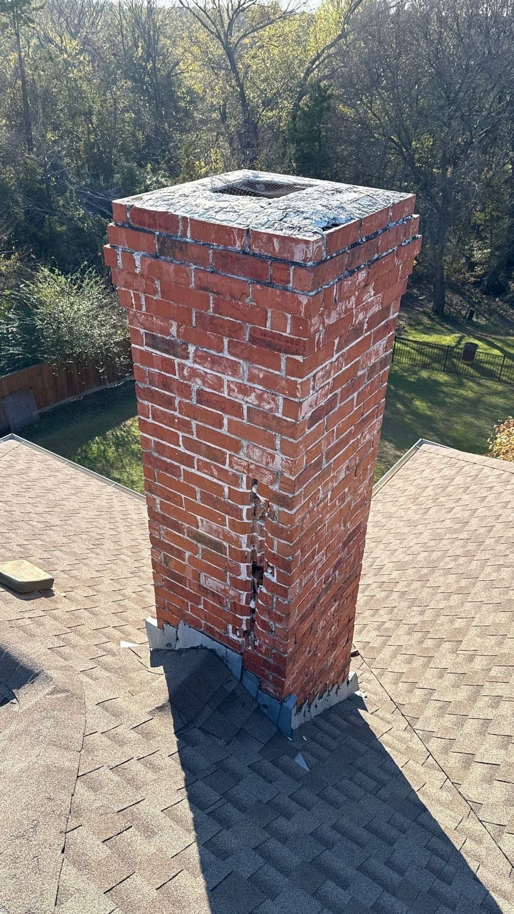 Brick chimney on rooftop with visible cracks and surrounding shingles, set against green trees.